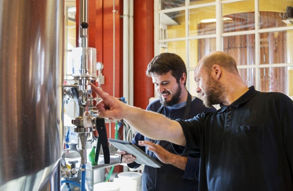 Two men looking at a tablet reviewing a vat of beer